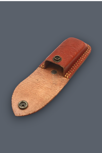 A small leather knife sheath or pouch made of two-toned brown leather with brass snap buttons, shown on a plain dark gray background.