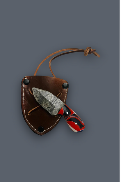A fixed-blade knife with a patterned silver blade and a red, black, and white handle, shown partially inserted into a brown leather sheath with a cord, on a solid dark gray background.
