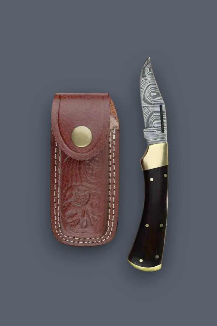 A folding pocket knife with a patterned steel blade, dark wood handle, and brass accents, displayed next to its brown leather sheath on a dark gray background.