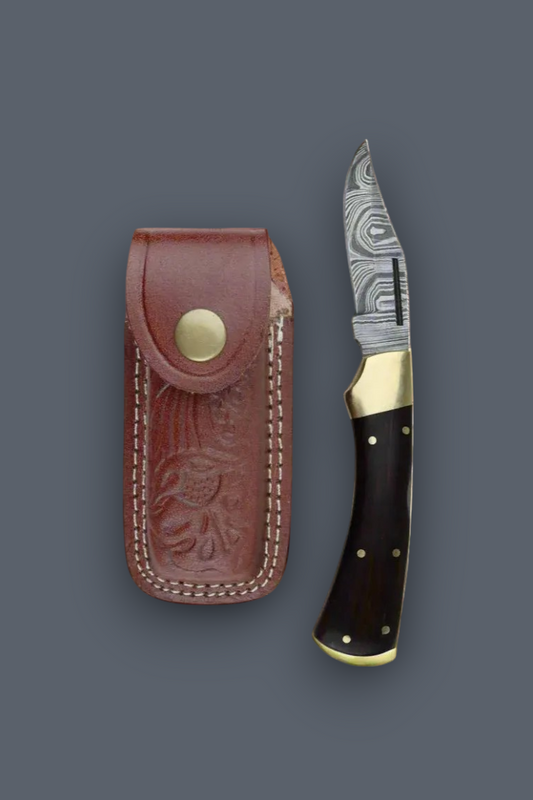 A folding pocket knife with a patterned steel blade, dark wood handle, and brass accents, displayed next to its brown leather sheath on a dark gray background.