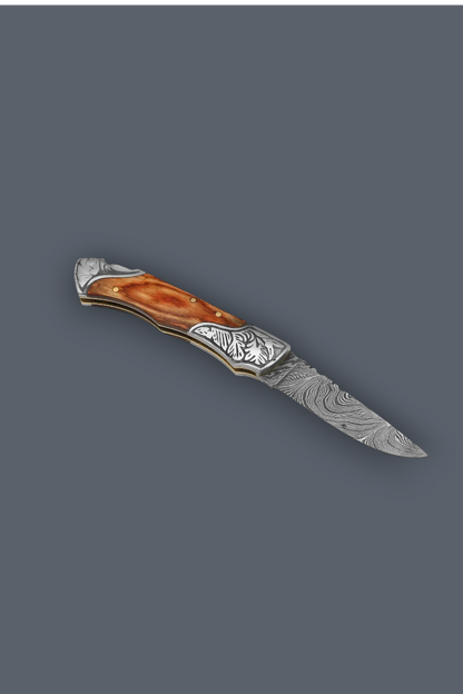 A folding pocket knife with a patterned Damascus steel blade, engraved silver-toned metal end caps, and a curved amber-brown handle inlay, shown on a solid dark gray background.