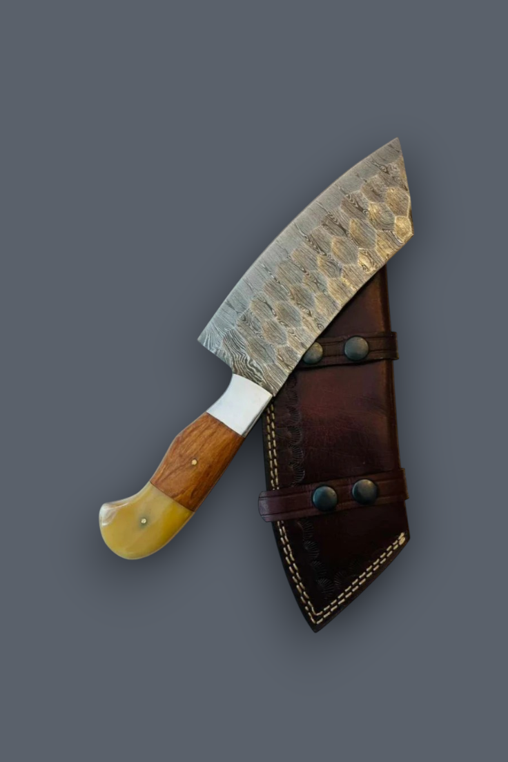 A handmade chef's knife with a hammered Damascus steel blade, a wooden handle, and a yellow bone bolster, shown next to its dark brown leather sheath on a solid gray background.