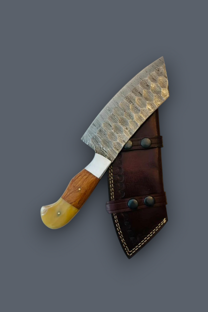 A handmade chef's knife with a hammered Damascus steel blade, a wooden handle, and a yellow bone bolster, shown next to its dark brown leather sheath on a solid gray background.