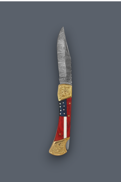 A custom folding pocket knife with a Damascus steel blade, gold-toned brass bolsters, and a handle featuring a red, white, and blue American flag design with brass pins, shown on a solid dark gray background.