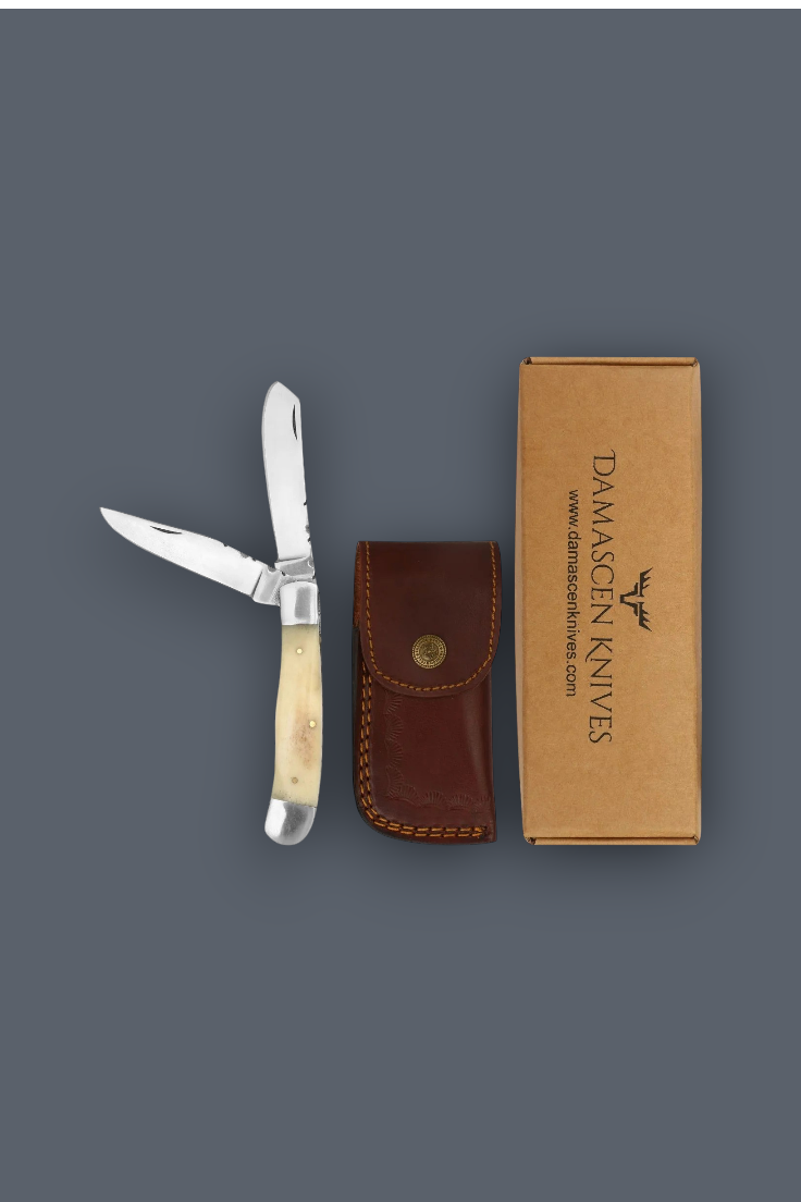Damason Knives folding pocket knife with an ivory bone handle, shown next to its brown leather sheath and a brown cardboard box on a dark gray background.