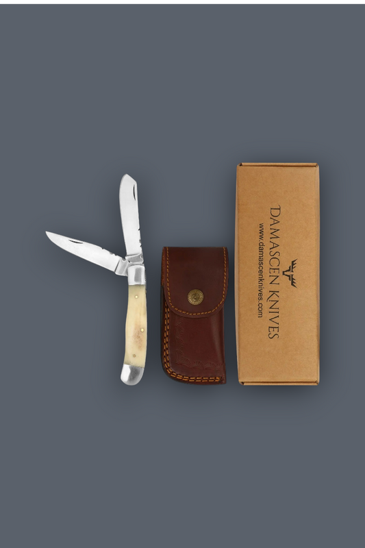 Damason Knives folding pocket knife with an ivory bone handle, shown next to its brown leather sheath and a brown cardboard box on a dark gray background.
