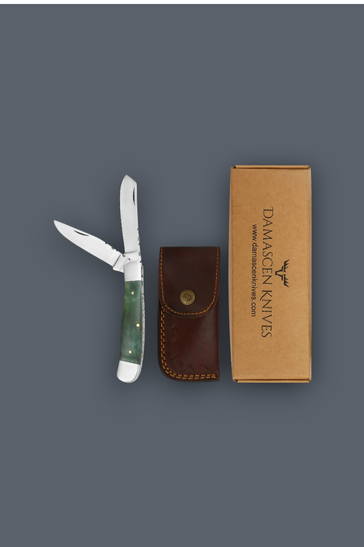 A folding pocket knife with a green handle is displayed next to its brown leather sheath and a brown cardboard box printed with the Damascen Knives logo, all arranged on a solid dark gray background.