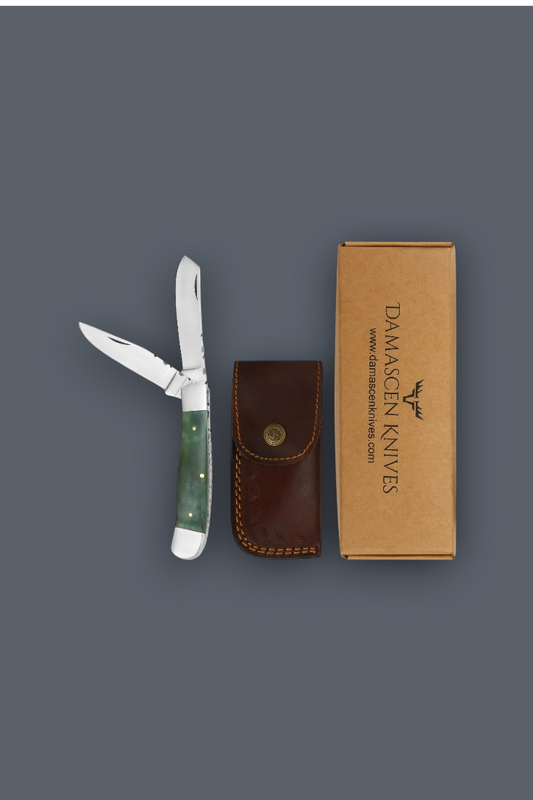 A folding pocket knife with a green handle is displayed next to its brown leather sheath and a brown cardboard box printed with the Damascen Knives logo, all arranged on a solid dark gray background.