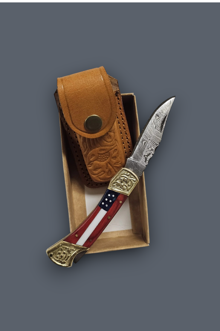 A folding pocket knife with a patterned steel blade, an American flag handle, and ornate brass-toned end caps, displayed next to its carved brown leather sheath inside an open presentation box on a dark gray background.