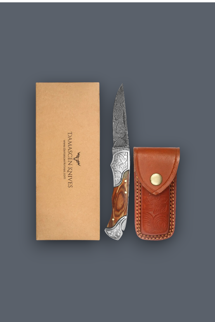 Damascus Knives folding pocket knife with a patterned steel blade and wood handle, shown next to its brown leather sheath and kraft paper packaging box on a dark gray background.