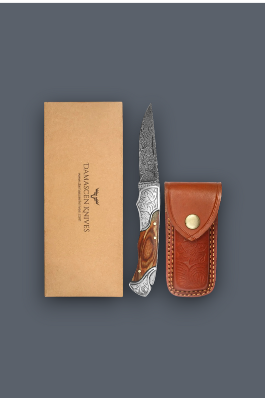 Damascus Knives folding pocket knife with a patterned steel blade and wood handle, shown next to its brown leather sheath and kraft paper packaging box on a dark gray background.