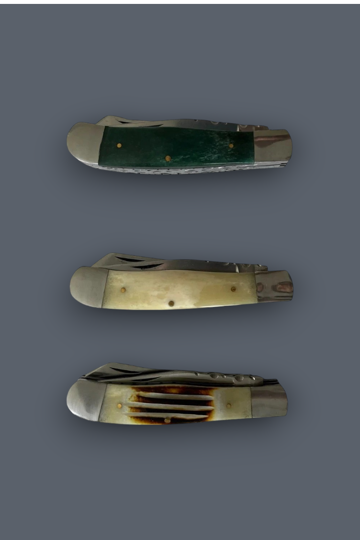 Three vintage folding pocket knives with bone handles are arranged vertically on a solid dark gray background. The top knife features a dark green bone inlay, the middle knife has cream-colored bone, and the bottom knife displays a natural gradient bone handle with dark burn marks.