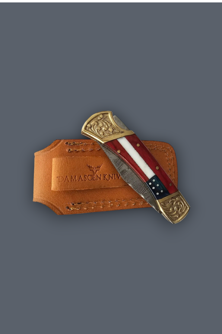 A folding pocket knife with a red, white, and blue striped handle and ornate gold-toned end caps, resting on a tan leather sheath embossed with 'DAMASCEN KNIFE' against a dark gray background.