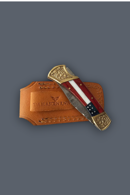 A folding pocket knife with a red, white, and blue striped handle and ornate gold-toned end caps, resting on a tan leather sheath embossed with 'DAMASCEN KNIFE' against a dark gray background.