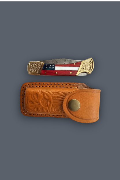 A folding pocket knife with a red, white, and blue flag inlay and ornate brass-toned ends, displayed above its matching tooled brown leather sheath on a dark gray background.