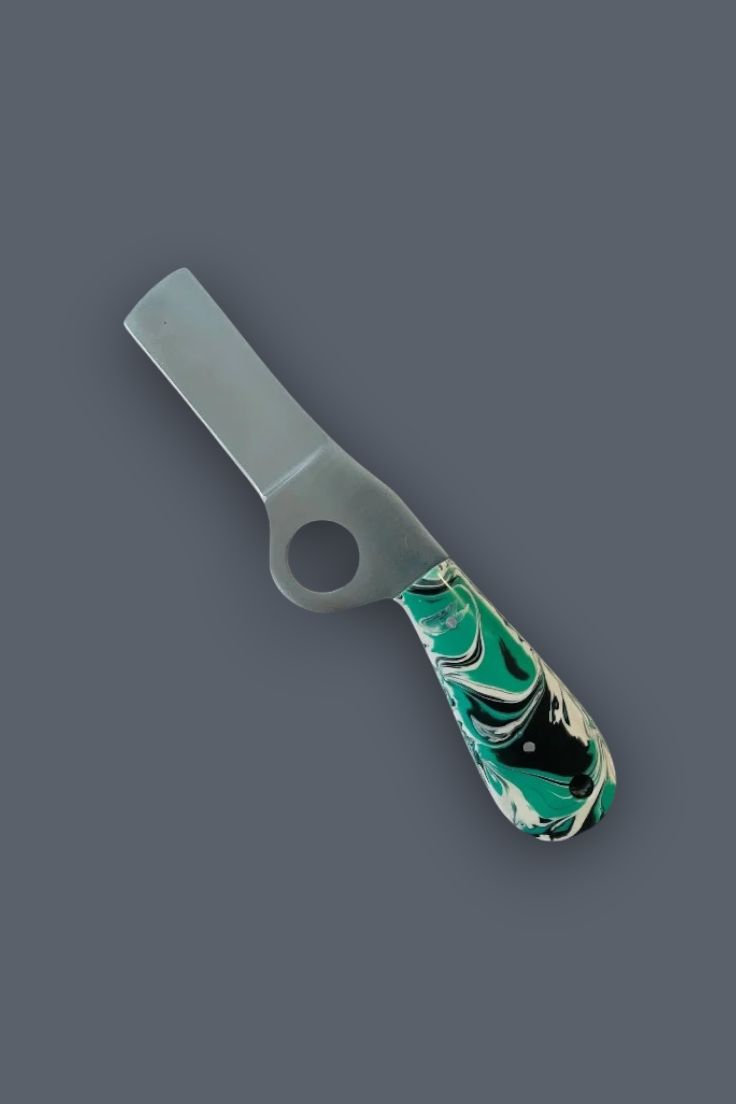 A small kitchen tool with a flat rectangular blade, a circular hole near the base, and a marbled green, black, and white handle, shown on a plain dark gray background.