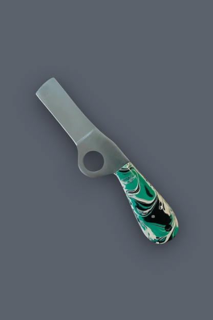 A small kitchen tool with a flat rectangular blade, a circular hole near the base, and a marbled green, black, and white handle, shown on a plain dark gray background.