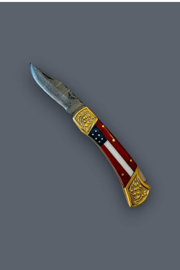 A folding pocket knife with a silver steel blade, ornate gold-toned metal accents, and a handle featuring a red, white, and blue American flag design, shown on a solid dark gray background.