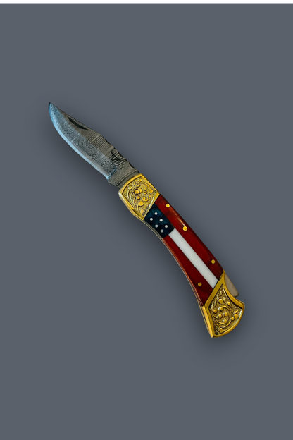 A folding pocket knife with a silver steel blade, ornate gold-toned metal accents, and a handle featuring a red, white, and blue American flag design, shown on a solid dark gray background.
