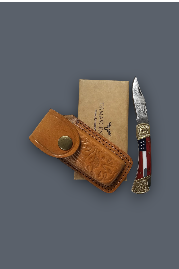 A folding pocket knife with a red, white, and blue striped handle and ornate gold-toned end pieces, displayed next to its tan leather sheath and a tan paper box printed with the Damace logo, all on a solid dark gray background.