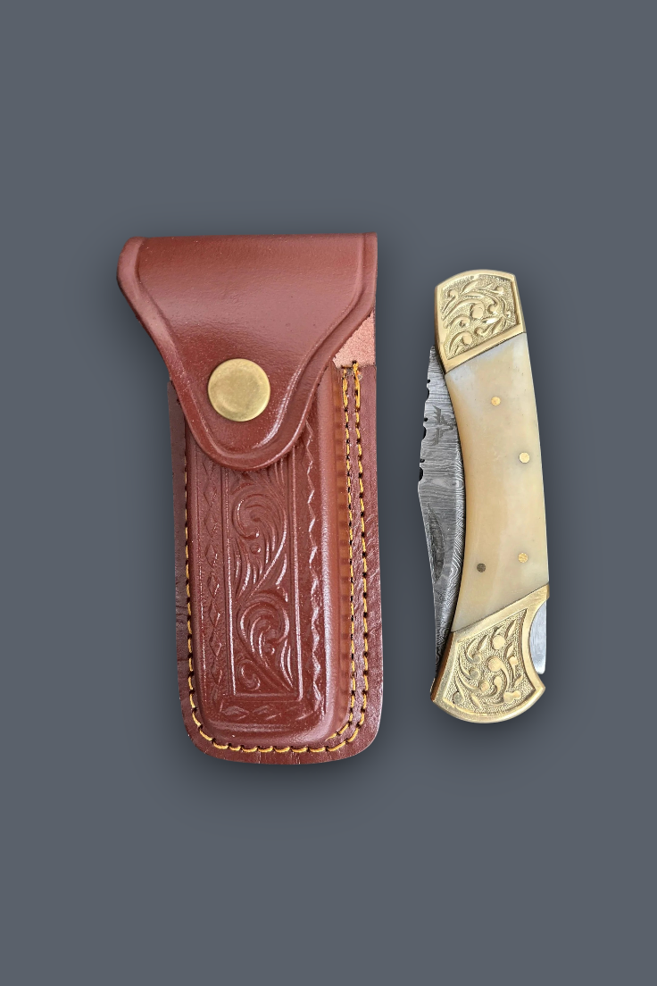 A folding pocket knife with an engraved bone or horn handle and brass accents, shown next to its brown leather sheath with floral tooling and a brass snap button, on a solid dark gray background.
