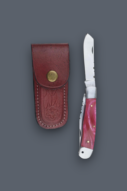 A folding pocket knife with a pink marbled handle and silver blade, shown next to its reddish-brown leather sheath with a brass snap button, on a dark gray background.