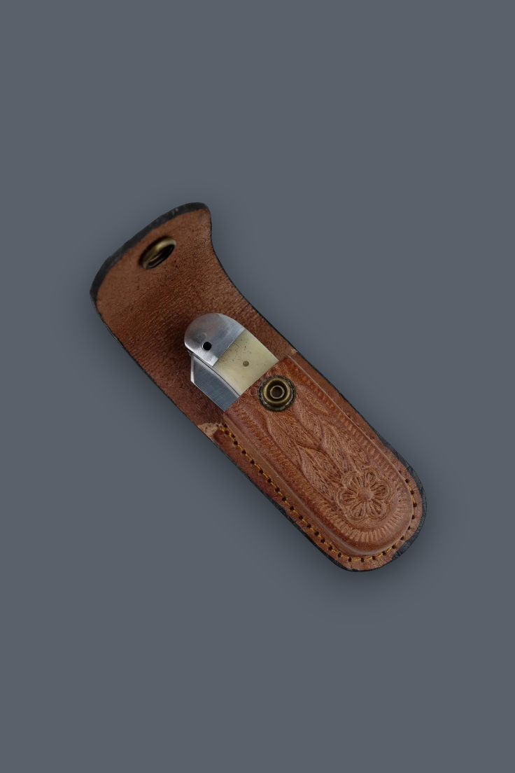 A brown leather knife sheath with floral tooling, holding a folding knife with a brass-colored handle, set against a solid dark gray background.