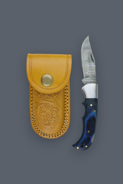 A folding pocket knife with a patterned steel blade and a dark blue marbled handle, shown next to its tan leather sheath on a solid dark gray background.