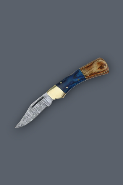 A folding pocket knife with a patterned steel blade, a polished brass bolster, and a handle made of blue marbled resin and natural wood, shown on a solid dark gray background.