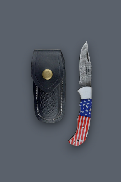 A compact folding pocket knife with sheath with a red, white, and blue American flag pattern handle, shown next to its black leather sheath with a brass snap button, on a solid dark gray background.