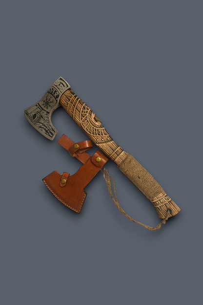 A Viking-style bearded axe with a carved wooden handle and engraved steel head, shown alongside a brown leather sheath on a solid dark gray background.