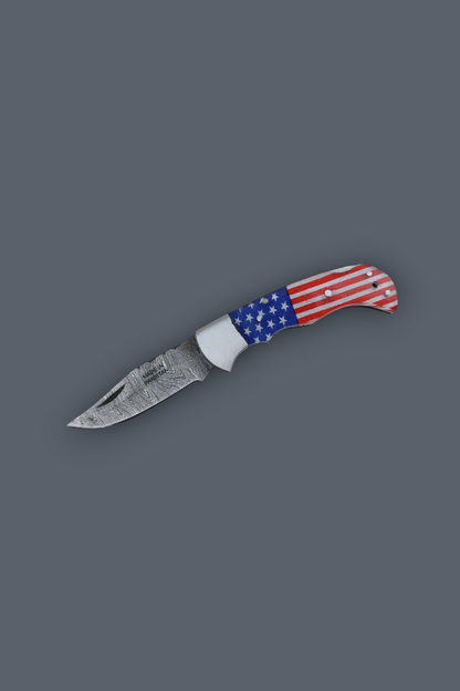 A pocket knife with a patterned steel blade and a handle featuring an American flag design, shown on a solid dark gray background.