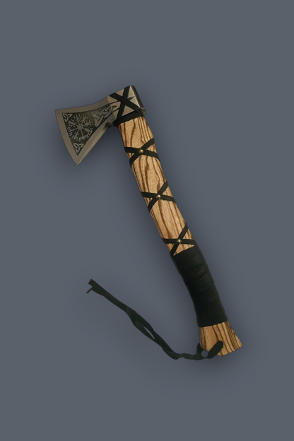 A decorative hand axe or hatchet with a carved light wood handle wrapped in black cord, a silver-toned metal head with an engraved geometric pattern, and a black wrist strap, shown on a solid dark gray background.