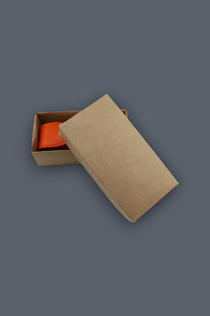 A brown kraft paper gift box with its lid propped open, revealing a bright orange item inside, set against a solid dark gray background.