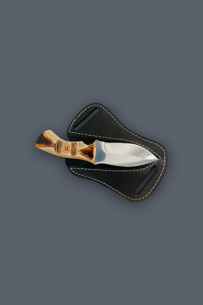 A small fixed-blade knife with a polished silver blade and a gold-toned handle featuring a red cross emblem, resting on a black leather sheath against a solid dark gray background.
