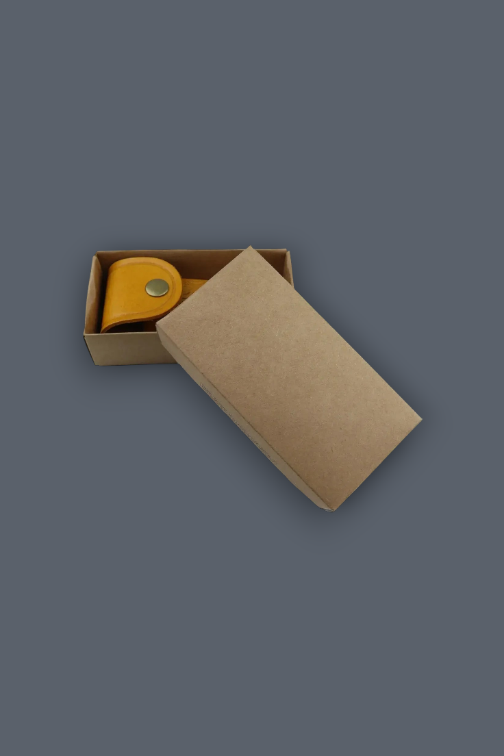A small yellow leather strap or accessory inside an open brown kraft paper box, with the box lid leaning against it, on a plain dark gray background.