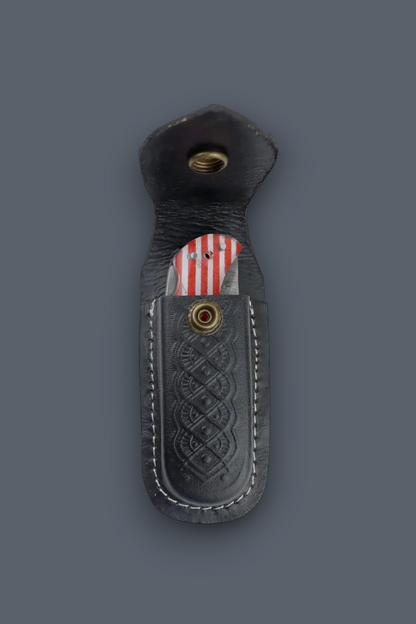 A black leather knife sheath with a stitched Celtic knot pattern and brass snap button, shown on a dark gray background with a knife partially inserted.