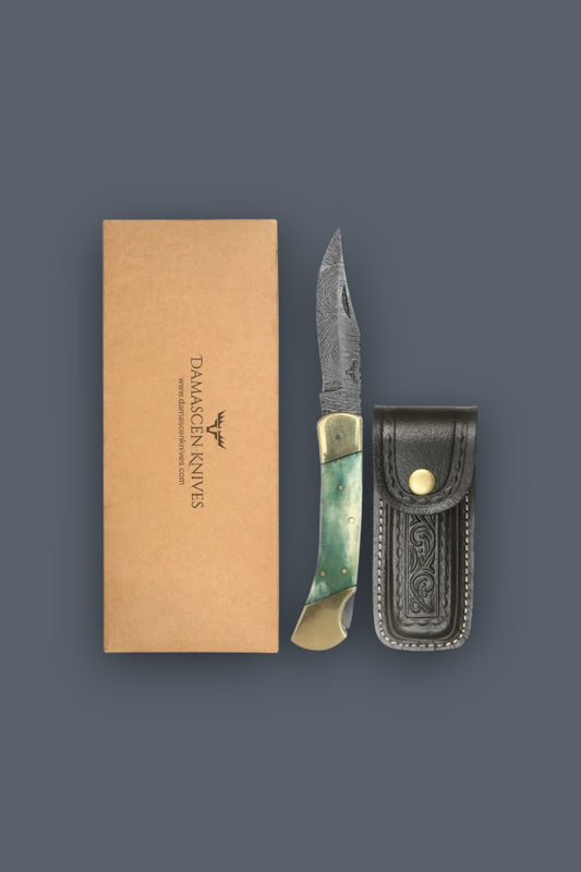 A folding pocket knife with a green and gold handle, displayed next to its brown cardboard packaging box and a black leather sheath on a dark gray background. The box is printed with the brand name 'DAARCEN KNIVES'.