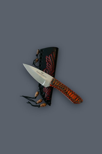 A fixed-blade knife with a silver blade and an orange-brown patterned handle, displayed diagonally in front of a black sheath with red designs and decorative beads, set against a solid dark gray background.
