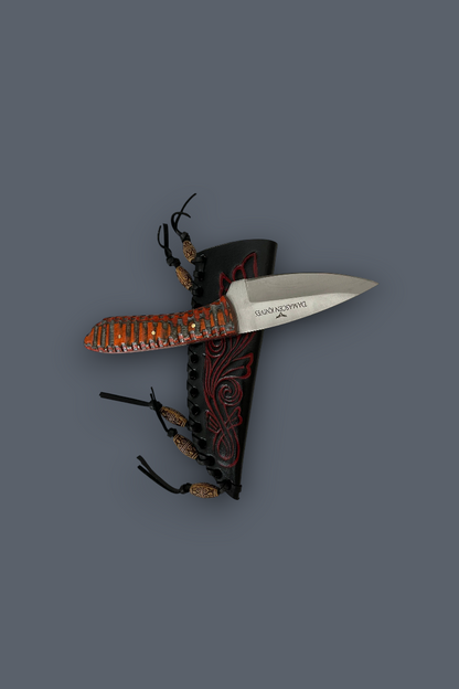 A fixed-blade knife with a silver blade and a textured orange-brown handle, displayed alongside a black sheath with red tribal-style patterns and black cords, set against a solid dark gray background.