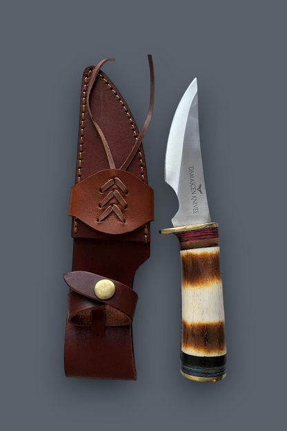 A fixed-blade knife with a polished silver blade and a carved bone handle, displayed next to its brown leather sheath on a dark gray background. The blade is engraved with the brand name 'Dakota Knife'.