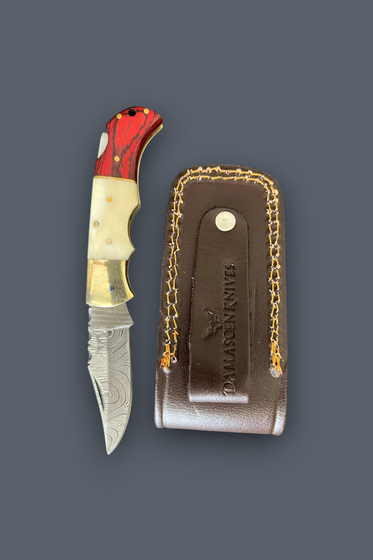 A folding pocket knife with a red and white marbled handle, gold-toned bolster, and patterned steel blade, shown next to its brown leather sheath with gold chain detailing on a dark gray background. The sheath is embossed with the brand name 'D.