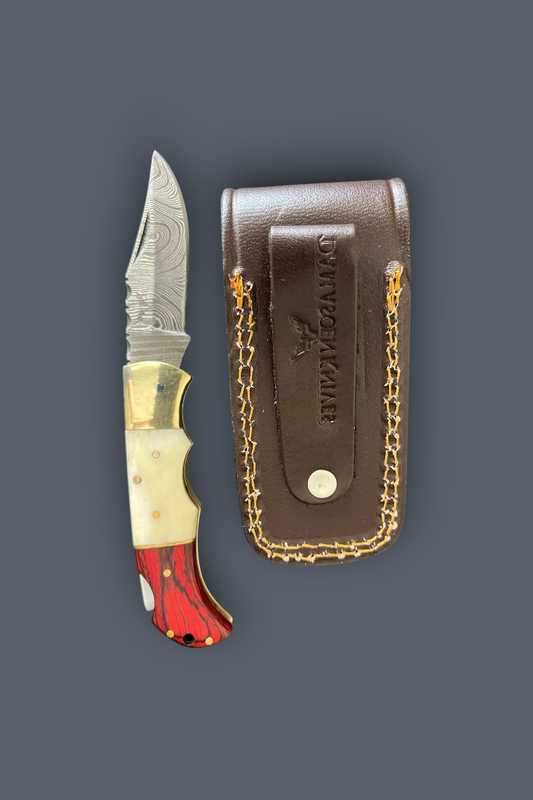 A folding pocket knife with a red and white marbled handle, gold-toned bolster, and patterned steel blade, shown next to its brown leather sheath with gold chain detailing on a dark gray background. The sheath is embossed with the brand name 'D.
