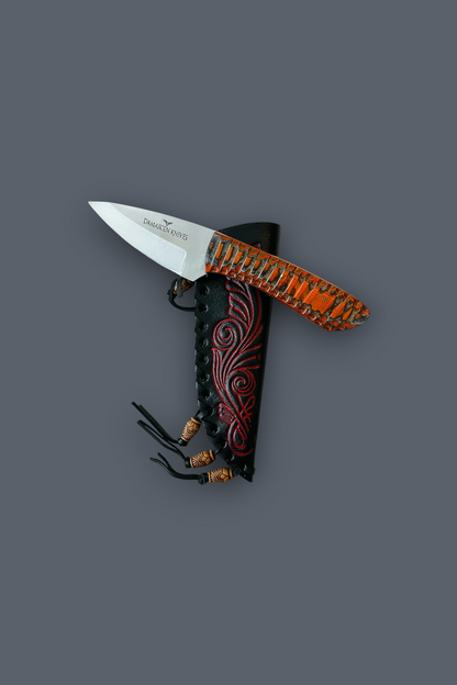 A fixed-blade knife with a silver blade and an orange-brown textured handle, shown partially sheathed in a black sheath with red scrollwork and black cords, on a solid dark gray background.