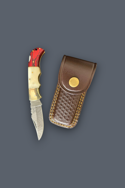 A folding pocket knife with a red and gold handle next to its brown leather sheath with gold-toned stitching, set against a solid dark gray background.