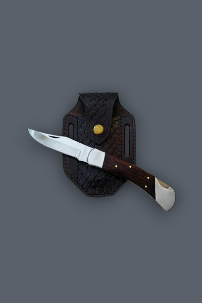 A folding pocket knife with a silver blade and dark brown wooden handle, displayed diagonally across a dark brown textured leather sheath on a solid dark gray background.