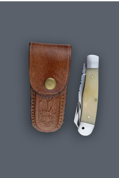 A folding pocket knife with a cream-colored bone handle and silver blade, displayed next to its brown leather sheath with an embossed eagle design and brass snap closure, set against a solid dark gray background.