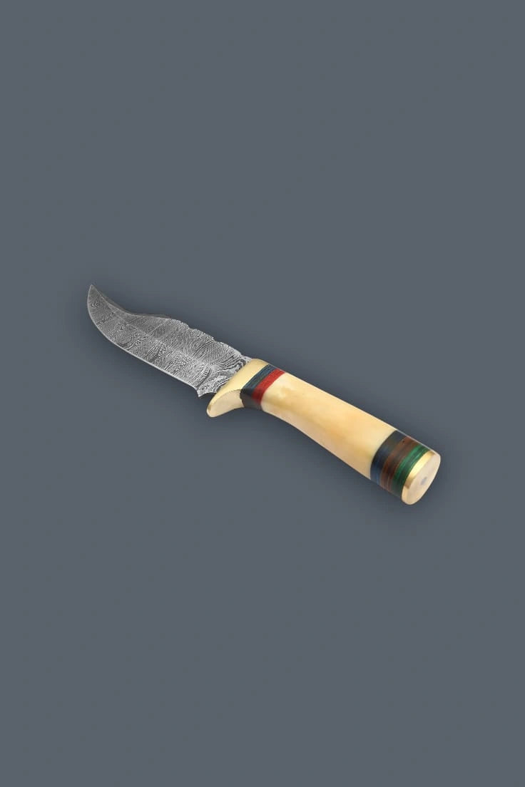 Downward diagonal flat lay of the Damascen Knives fixed blade, emphasizing the polished brass pommel, ergonomic bone grip, and multi-colored handle spacers.