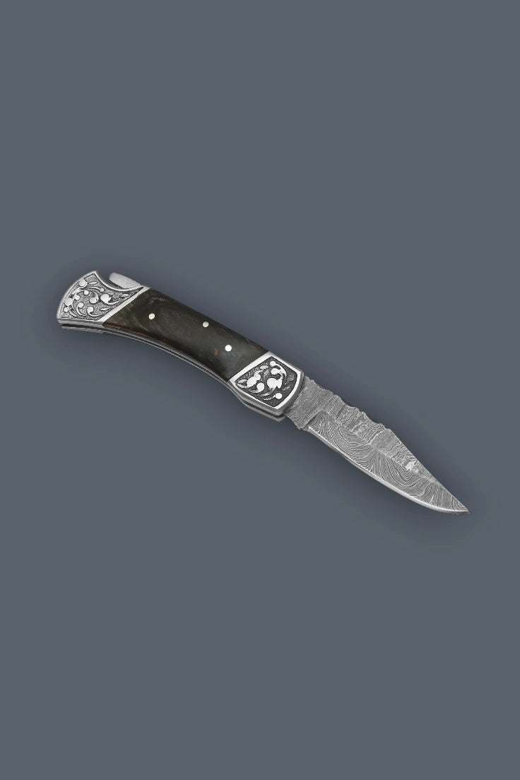 Angled flat lay of the fully open The Squire Damascus pocket knife showcasing the ornate engraved bolsters and multicolor wood handle.