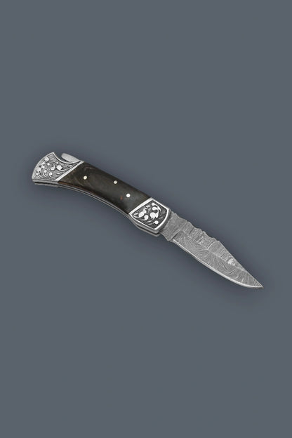 Angled flat lay of the fully open The Squire Damascus pocket knife showcasing the ornate engraved bolsters and multicolor wood handle.